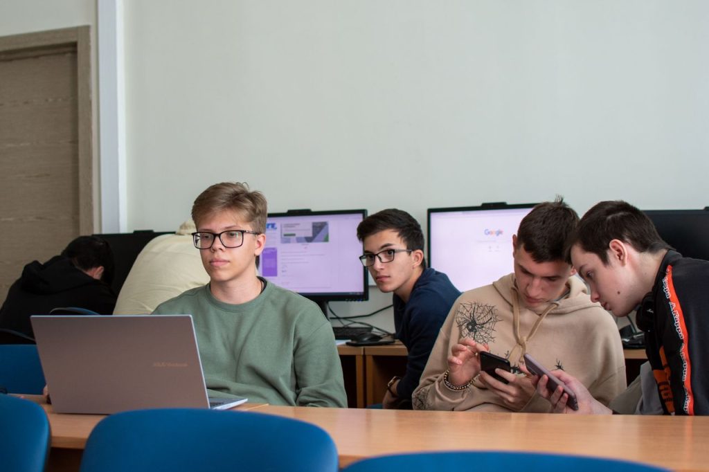 Five teenagers engaged with laptops and smartphones in a modern classroom.